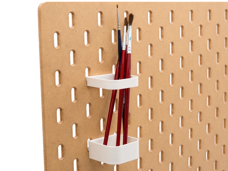 May include: A light brown pegboard with evenly spaced holes. Two white, square-shaped holders are attached, each holding several red paint brushes. The brushes have dark brown bristles and are of varying sizes.