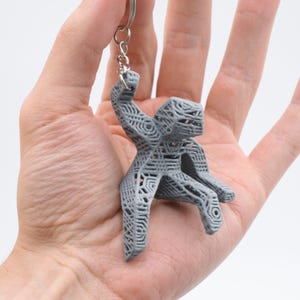 Puede incluir: Un llavero gris impreso en 3D con una figura humana estilizada sentada. La figura tiene un diseño abierto, similar a una celosía, con estructuras internas visibles. El llavero está unido a un anillo de metal plateado.