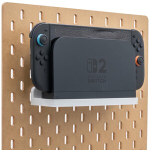 May include: A Nintendo Switch console and dock are mounted on a brown pegboard. The Switch is black with blue and orange accents. The dock is dark grey with the Nintendo Switch logo and sits on a white shelf.