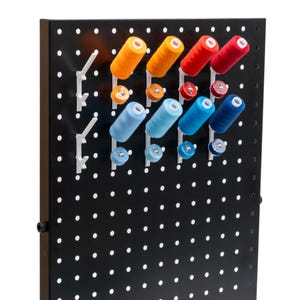 May include: A black pegboard with white pegs holding spools of thread. The thread spools are orange, red, and various shades of blue. The bobbins are in matching colors. This sewing organizer is perfect for storing thread and bobbins.