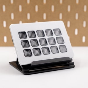 May include: A small, rectangular, electronic device with a grid of 16 square, translucent buttons. The device is white with a black base, and is designed to stand upright. The buttons appear to be slightly raised and have a textured surface.