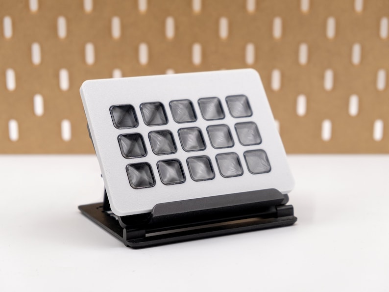 May include: A small, rectangular, electronic device with a grid of 16 square, translucent buttons. The device is white with a black base, and is designed to stand upright. The buttons appear to be slightly raised and have a textured surface.