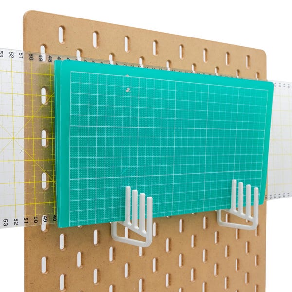 May include: A brown pegboard with a green cutting mat and clear rulers. The cutting mat is held in place by white plastic brackets. The rulers are marked with inch measurements.