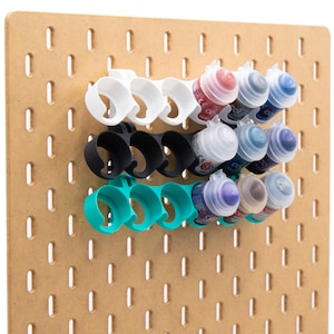 May include: A light brown pegboard with three rows of paint bottle holders in white, black, and teal. Each holder securely cradles several paint bottles in various colours, including red, blue, and purple. The pegboard is made of wood.