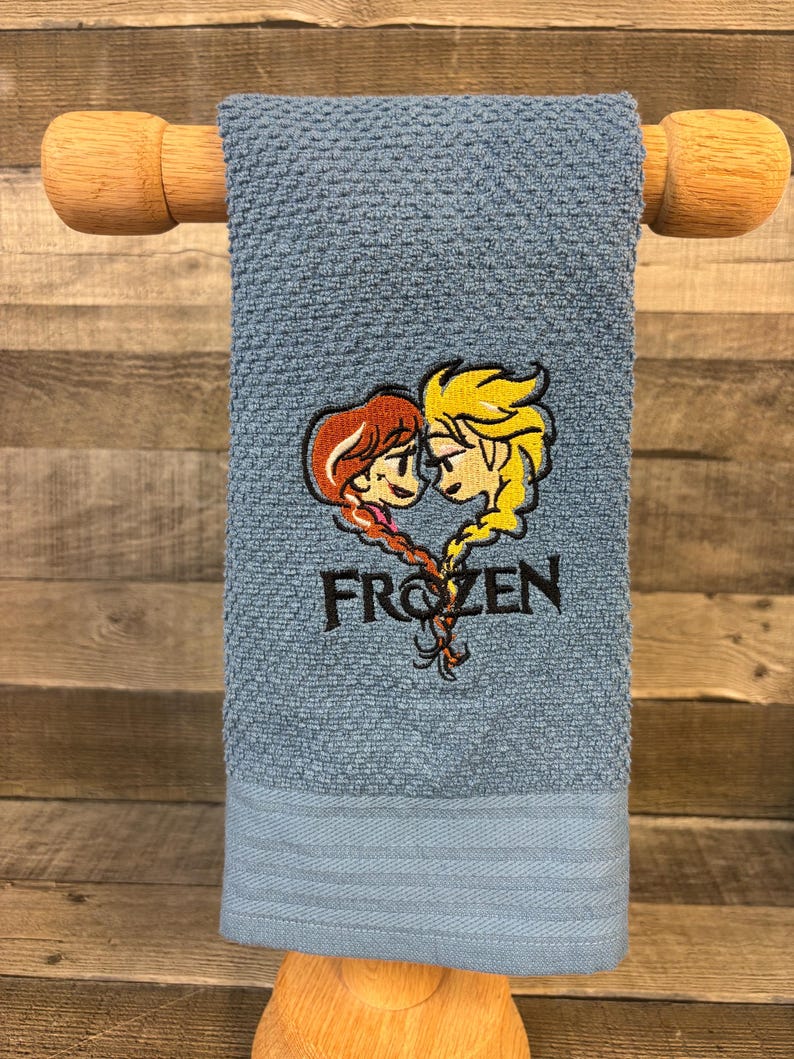May include: A blue textured hand towel featuring an embroidered design of two characters with braided hair forming a heart shape above the black text FROZEN. The towel is draped over a wooden rod and has a smooth hemmed border at the bottom edge.