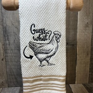 May include: White kitchen towel with brown stripes at the bottom. The towel has a black embroidered image of a chicken with the text "Guess what?" above it.