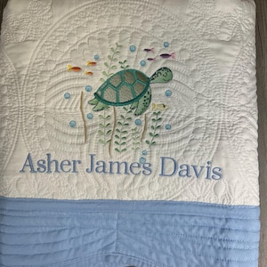 May include: White quilted baby blanket with an embroidered sea turtle design in shades of green and blue. The blanket features the embroidered name "Asher James Davis" in blue thread. The bottom border is a light blue fabric with horizontal stitching.