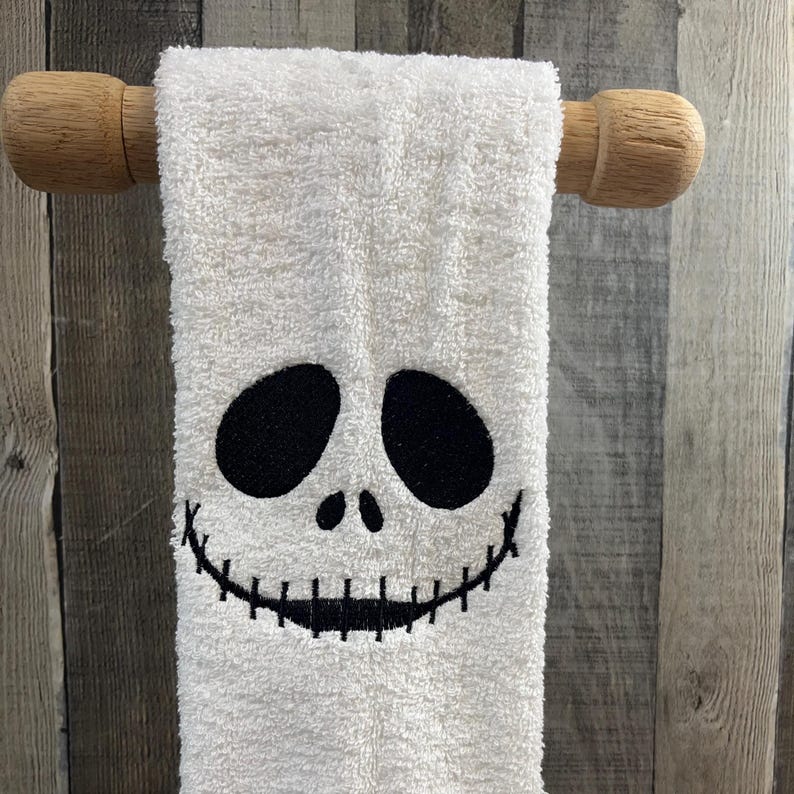 K&ouml;nnte beinhalten: Wei&szlig;es Handtuch mit einem schwarzen, gestickten Gesicht, das an eine Halloween-Figur erinnert. Das Handtuch h&auml;ngt an einer Holzstange. Das Gesicht hat gro&szlig;e ovale Augen und ein gesticktes L&auml;cheln.