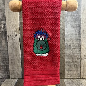May include: Red kitchen towel with an embroidered green monster wearing a red hat. The monster has blue hair and big eyes.