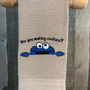 May include: Tan hand towel with the embroidered image of a blue monster peeking over the bottom edge. The text "Are you making cookies?" is above the monster. The towel is hanging on a wooden towel rack.