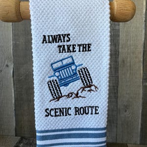 Puede incluir: Una toalla de mano blanca con tejido de gofre que presenta un veh&iacute;culo todoterreno azul bordado sobre un terreno rocoso marr&oacute;n. El texto "ALWAYS TAKE THE SCENIC ROUTE" est&aacute; bordado en hilo negro sobre y bajo el veh&iacute;culo. Franjas azules en la base.