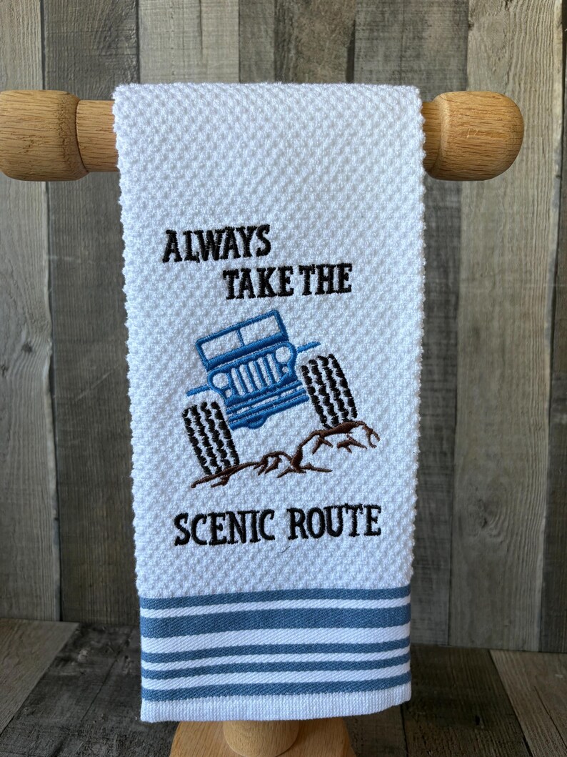 Puede incluir: Una toalla de mano blanca con tejido de gofre que presenta un veh&iacute;culo todoterreno azul bordado sobre un terreno rocoso marr&oacute;n. El texto "ALWAYS TAKE THE SCENIC ROUTE" est&aacute; bordado en hilo negro sobre y bajo el veh&iacute;culo. Franjas azules en la base.