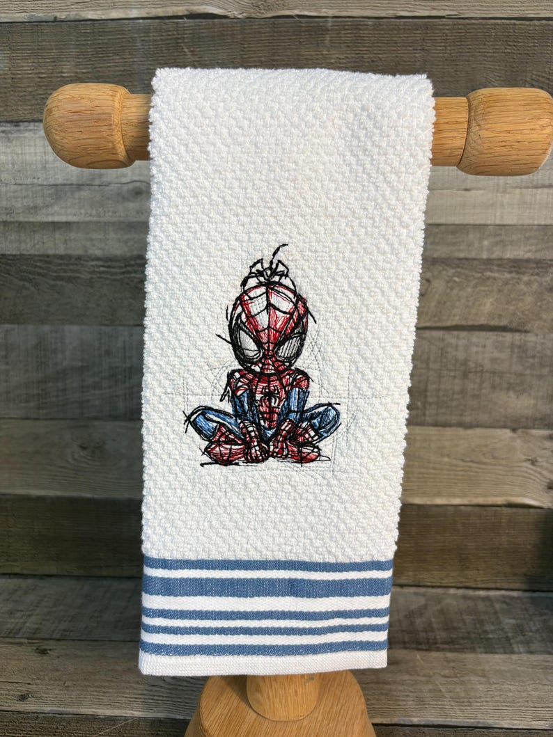 May include: White hand towel with a textured weave, featuring an embroidered Spider-Man design in red, blue, and black. The towel has blue and white striped accents at the bottom and is displayed on a wooden towel rack.