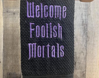 以下が含まれることがあります： 「Welcome Foolish Mortals」という紫色の刺繍が施された黒いキッチンタオルです。