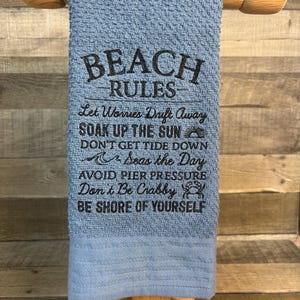 May include: A blue beach towel with the words "BEACH RULES" embroidered in black. The towel includes phrases like "Let Worries Drift Away" and "Soak Up The Sun". The towel is displayed on a wooden towel rack.