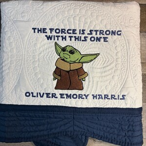 Puede incluir: Manta acolchada blanca con borde azul marino. La manta presenta una imagen bordada de un personaje alien&iacute;gena verde con el texto "THE FORCE IS STRONG WITH THIS ONE" y un nombre.