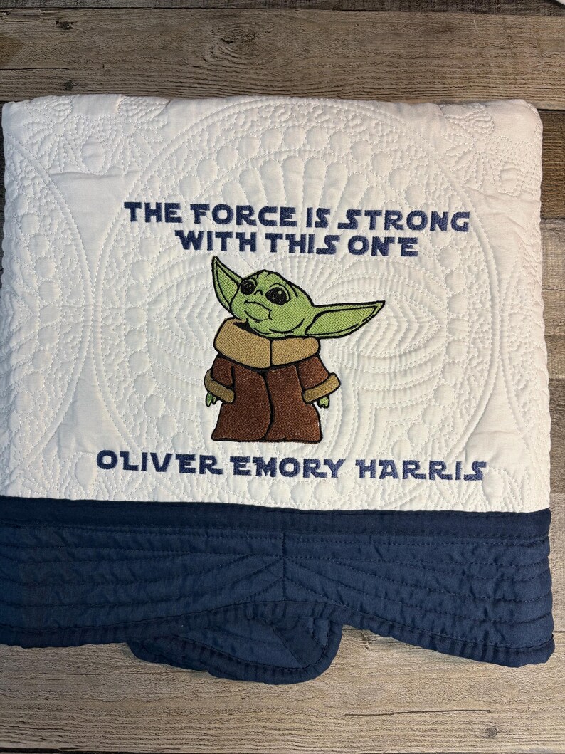 Puede incluir: Manta acolchada blanca con borde azul marino. La manta presenta una imagen bordada de un personaje alien&iacute;gena verde con el texto "THE FORCE IS STRONG WITH THIS ONE" y un nombre.