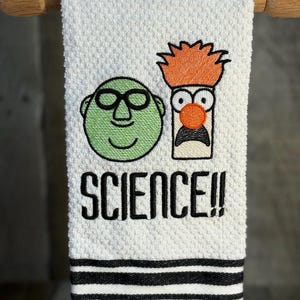 May include: White hand towel with black and white stripes at the bottom. The towel features embroidered cartoon characters and the word "SCIENCE!!" in black. The characters include a green face with glasses and a character with orange hair.