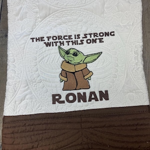 Puede incluir: Manta de beb&eacute; blanca con borde marr&oacute;n y la frase bordada "THE FORCE IS STRONG WITH THIS ONE" y el nombre "RONAN". La manta presenta una imagen bordada de un personaje alien&iacute;gena verde.