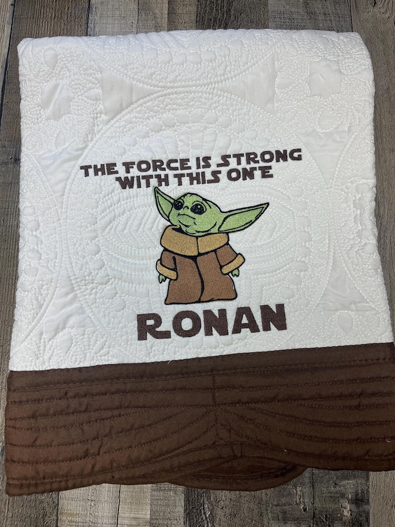 Puede incluir: Manta de beb&eacute; blanca con borde marr&oacute;n y la frase bordada "THE FORCE IS STRONG WITH THIS ONE" y el nombre "RONAN". La manta presenta una imagen bordada de un personaje alien&iacute;gena verde.