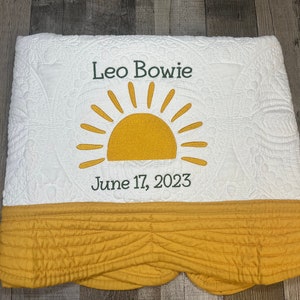 May include: White quilted blanket with a yellow sun design and the name "Leo Bowie" embroidered in green. The date "June 17, 2023" is also embroidered. The bottom edge features a yellow, scalloped border.