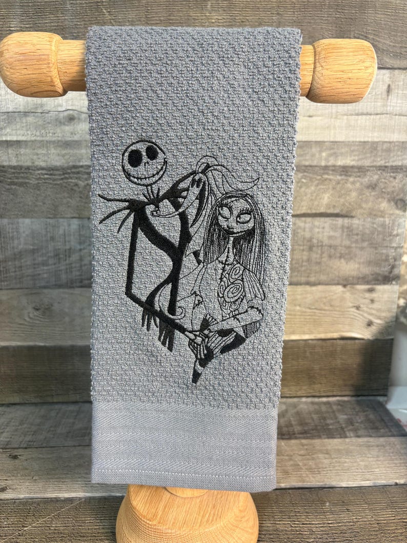 May include: Gray hand towel featuring an embroidered design of Jack Skellington, Sally, and Zero from The Nightmare Before Christmas. The towel is displayed on a wooden towel rack.