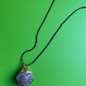 Könnte beinhalten: Ein Anhänger aus violettem Amethystkristall an einer schwarzen Kettenkette. Der Kristall ist roh und hat eine natürliche, unregelmäßige Form.