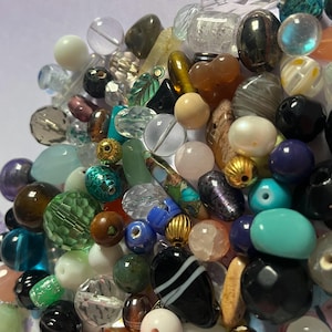 Op de afbeelding: Een verzameling diverse kralen in verschillende vormen en kleuren. De kralen omvatten heldere, ondoorzichtige en iriserende varianten, sommige met metallic accenten. De collectie bevat ronde, ovale en onregelmatige vormen, geschikt voor het maken van sieraden of knutselen.