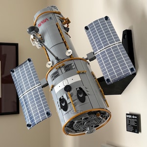 Puede incluir: Un modelo LEGO del telescopio espacial Hubble de la NASA montado en una pared. El cuerpo cilíndrico gris presenta detalles dorados, dos grandes paneles solares con patrón de rejilla azul y blanco, y el logotipo de la NASA. Una placa negra está al lado.