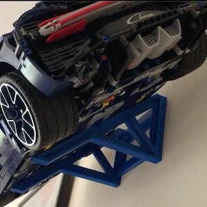 May include: A blue Lego Bugatti Chiron model car displayed on a blue wall mount. The car is upside down with the rear end facing the camera. The car has a black and silver engine and a black spoiler. The wall mount is a triangular shape with a blue finish.
