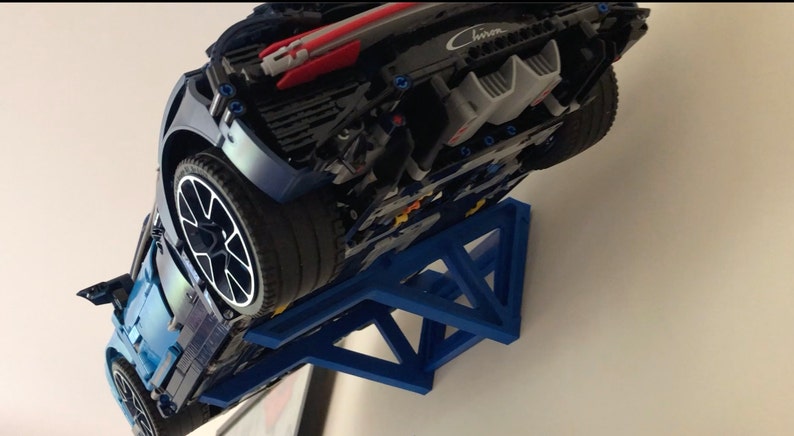 May include: A blue Lego Bugatti Chiron model car displayed on a blue wall mount. The car is upside down with the rear end facing the camera. The car has a black and silver engine and a black spoiler. The wall mount is a triangular shape with a blue finish.