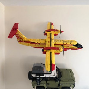 Op de afbeelding: Een geel en rood speelgoedvliegtuig met zwarte propellers en het woord "FIRE" erop gedrukt. Het vliegtuig is aan een muur gemonteerd met een groene speelgoedtruck eronder.