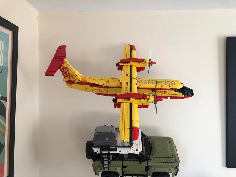 Op de afbeelding: Een geel en rood speelgoedvliegtuig met zwarte propellers en het woord "FIRE" erop gedrukt. Het vliegtuig is aan een muur gemonteerd met een groene speelgoedtruck eronder.