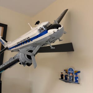 Può includere: Un modello di aereo Pan American bianco e blu con eliche rotanti. Il modello è montato su una parete, con un piccolo logo Pan American e figurine sottostanti. Il modello ha le parole "PAN AMERICAN" stampate sul lato.