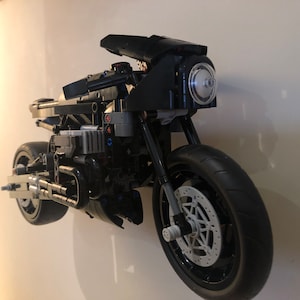 May include: A black Lego motorcycle model with a clear headlight and a detailed engine. The motorcycle is mounted on a white wall.