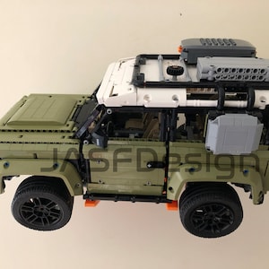 Op de afbeelding: Een groen Lego Land Rover Defender model met zwarte wielen en een wit dak. Het model heeft een dakrek met een grijze opbergbox en een zwarte reserveband.