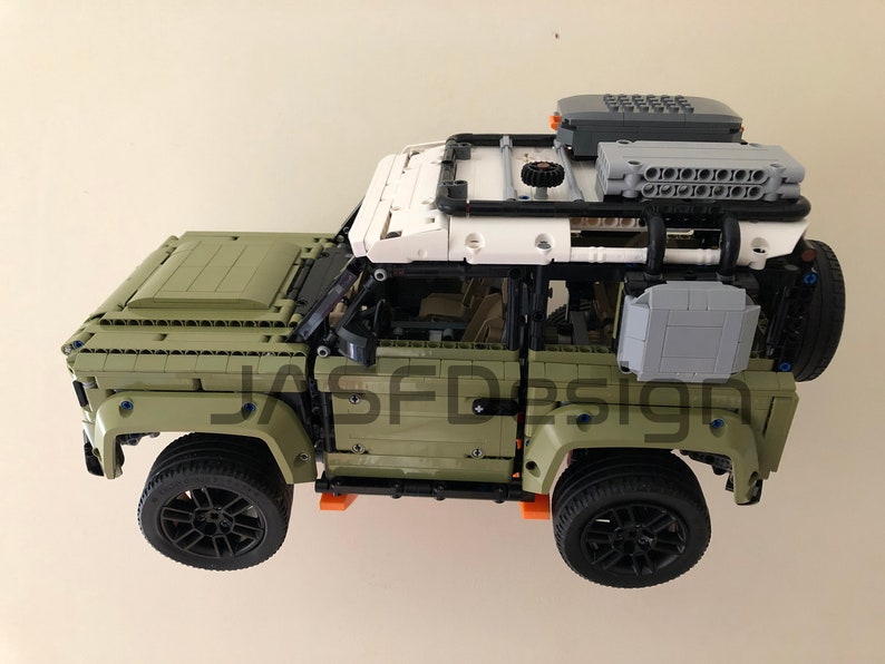 Op de afbeelding: Een groen Lego Land Rover Defender model met zwarte wielen en een wit dak. Het model heeft een dakrek met een grijze opbergbox en een zwarte reserveband.