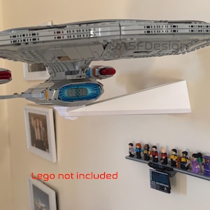 Puede incluir: Un modelo gris de la nave espacial Star Trek Enterprise montado en una estanter&iacute;a blanca. Debajo, una estanter&iacute;a muestra figuras de Star Trek. El texto "Lego not included" es visible en rojo. La estanter&iacute;a est&aacute; montada en una pared beige.