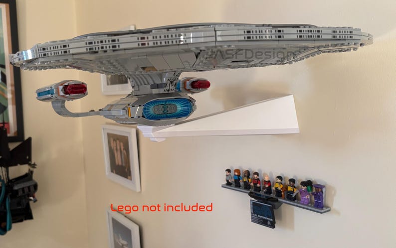 Puede incluir: Un modelo gris de la nave espacial Star Trek Enterprise montado en una estanter&iacute;a blanca. Debajo, una estanter&iacute;a muestra figuras de Star Trek. El texto "Lego not included" es visible en rojo. La estanter&iacute;a est&aacute; montada en una pared beige.