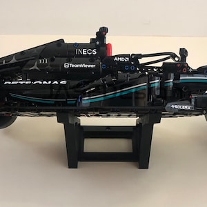 May include: A black and silver Lego model of a Formula 1 race car with the Petronas logo and Ineos branding. The car is mounted on a black stand.