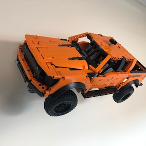 Op de afbeelding: Een oranje Lego Ford Raptor pick-up truck met zwarte wielen en een zwart interieur. De truck hangt in de lucht tegen een witte muur.