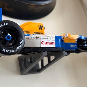 May include: A blue, yellow, and white race car model with the number 5 on the side. The car has black tires with "Goodyear" and "Eagle" branding. The word "Canon" is printed on the side. The model is mounted on a gray bracket.