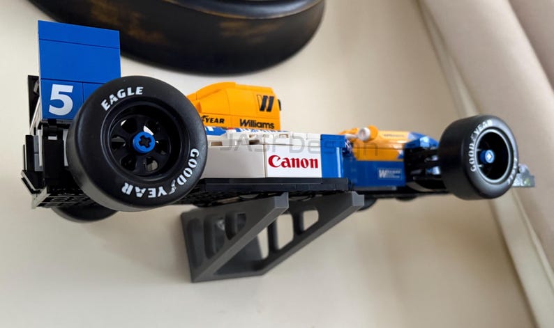 May include: A blue, yellow, and white race car model with the number 5 on the side. The car has black tires with "Goodyear" and "Eagle" branding. The word "Canon" is printed on the side. The model is mounted on a gray bracket.