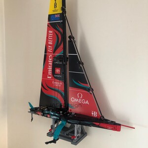 K&ouml;nnte beinhalten: Ein rotes, schwarzes und wei&szlig;es LEGO-Modell eines Segelboots mit den Worten "Emirates Fly Better" und "Omega" auf dem Segel. Das Modell ist auf einem grauen St&auml;nder montiert und hat ein blaues und t&uuml;rkisfarbenes Ruder.