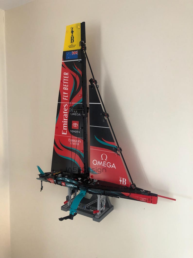 K&ouml;nnte beinhalten: Ein rotes, schwarzes und wei&szlig;es LEGO-Modell eines Segelboots mit den Worten "Emirates Fly Better" und "Omega" auf dem Segel. Das Modell ist auf einem grauen St&auml;nder montiert und hat ein blaues und t&uuml;rkisfarbenes Ruder.