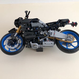 Peut inclure: Un mod&egrave;le de moto LEGO noir et gris avec des roues bleues et une cha&icirc;ne dor&eacute;e. La moto est suspendue en l'air contre un mur blanc.