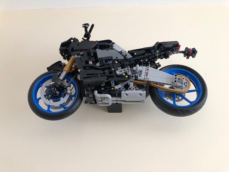 Peut inclure: Un mod&egrave;le de moto LEGO noir et gris avec des roues bleues et une cha&icirc;ne dor&eacute;e. La moto est suspendue en l'air contre un mur blanc.