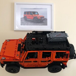 May include: An orange Lego model of a Mercedes-Benz G-Wagon with a black roof rack and a gray cargo box. The model is on a white surface and there is a framed photo of a real Mercedes-Benz G-Wagon in the background.