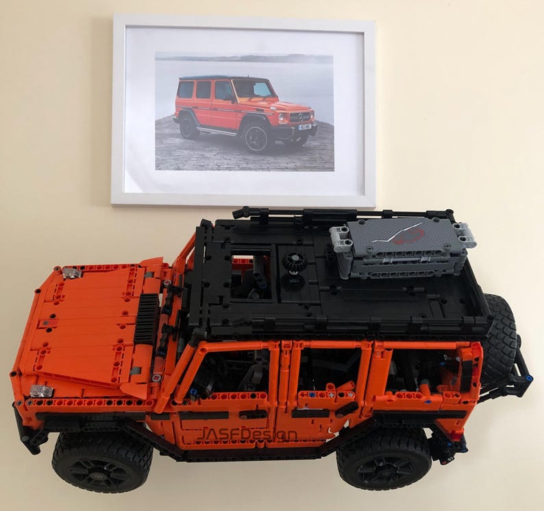May include: An orange Lego model of a Mercedes-Benz G-Wagon with a black roof rack and a gray cargo box. The model is on a white surface and there is a framed photo of a real Mercedes-Benz G-Wagon in the background.