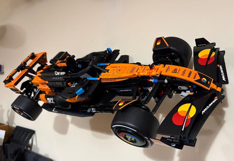 May include: A black and orange Lego McLaren Formula 1 race car model. The model features black tires, blue accents, and various sponsor logos. The car is mounted on a white wall.
